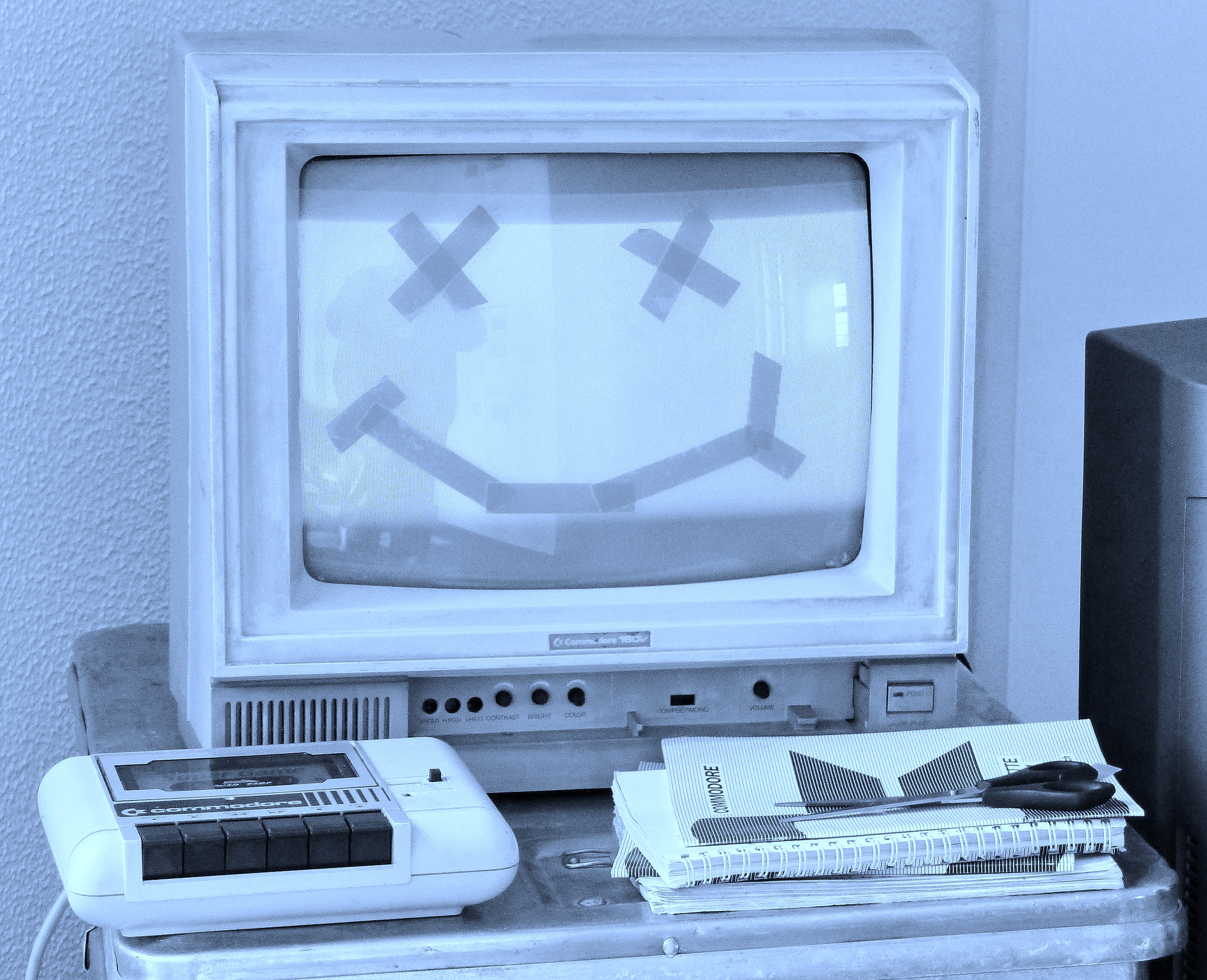 An old computer with a smiley face drawn in masking tape over the screen. An old tape recorder, a few notebooks, and a pair of scissors sit on a desk.